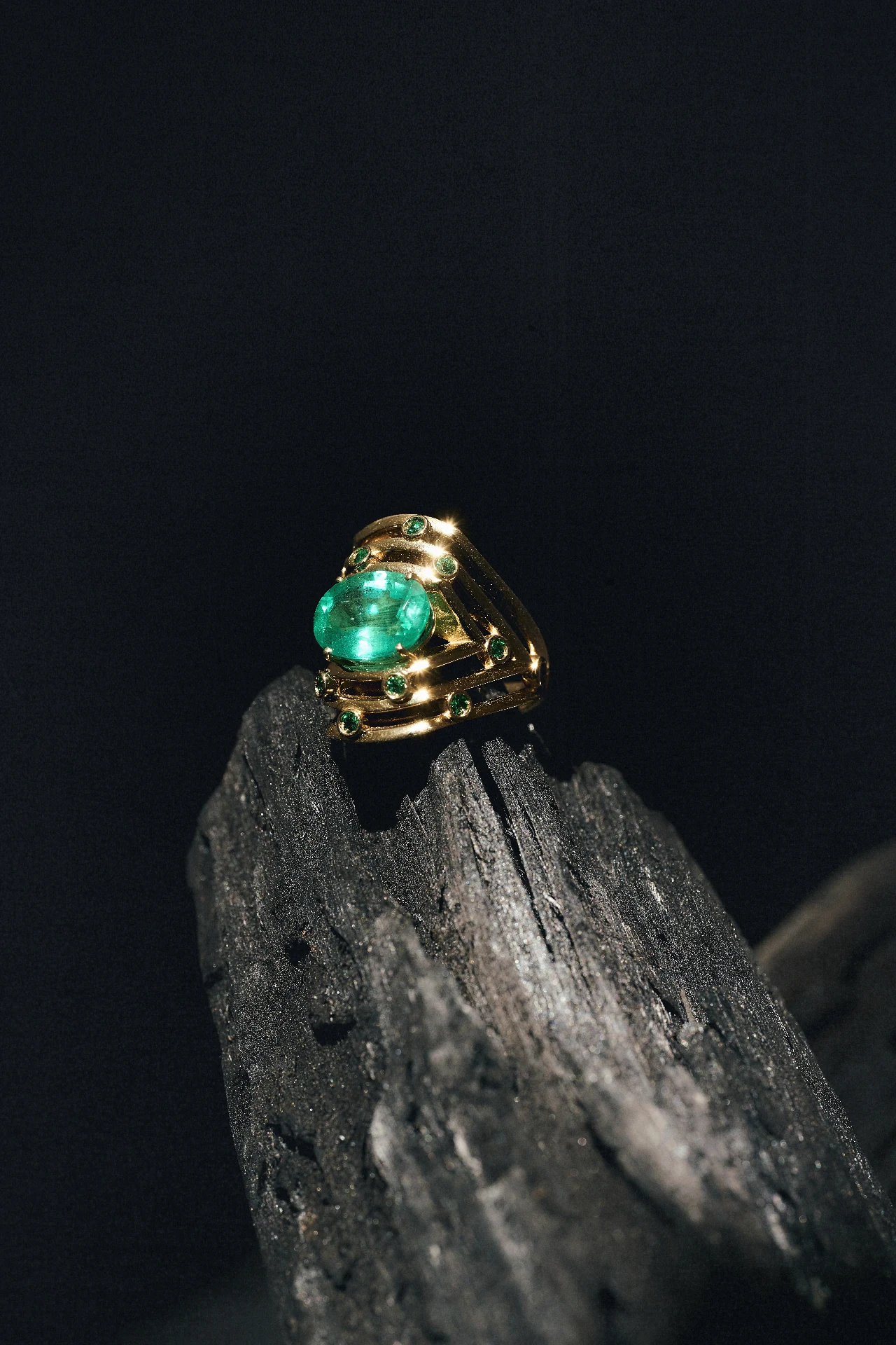 Anel ORIGEM em ouro 18k com esmeralda Belmont oval e esmeraldas acentuadas, sobre fragmento de carvão